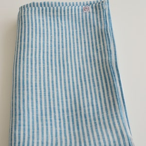 May include: Folded blue and white striped linen fabric. The fabric features vertical stripes of light blue and white. A small circular tag is visible on the top right corner of the folded fabric.