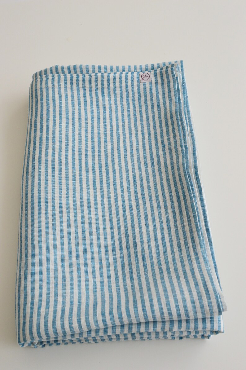 May include: Folded blue and white striped linen fabric. The fabric features vertical stripes of light blue and white. A small circular tag is visible on the top right corner of the folded fabric.