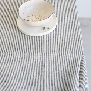 May include: A cream-colored ceramic bowl with numerous small holes, resting on a white plate. The bowl and plate are on a striped tablecloth with vertical gray and white stripes. A small sprig of greenery is on the plate.