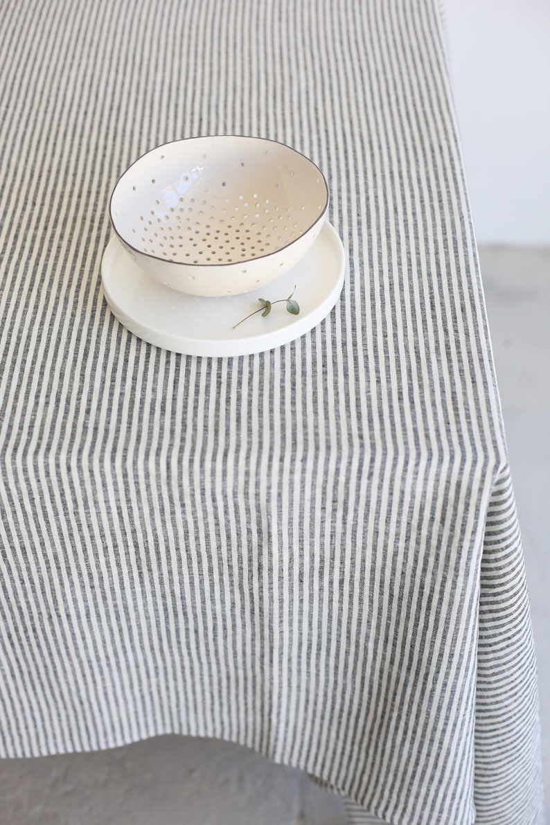 May include: A cream-colored ceramic bowl with numerous small holes, resting on a white plate. The bowl and plate are on a striped tablecloth with vertical gray and white stripes. A small sprig of greenery is on the plate.
