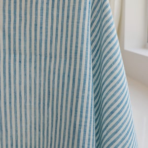May include: Close-up of a blue and white striped fabric. The fabric is draped, showing vertical stripes of varying widths. The material appears to be linen or a similar natural fiber, with a slightly textured surface. The color palette is a light blue and white.