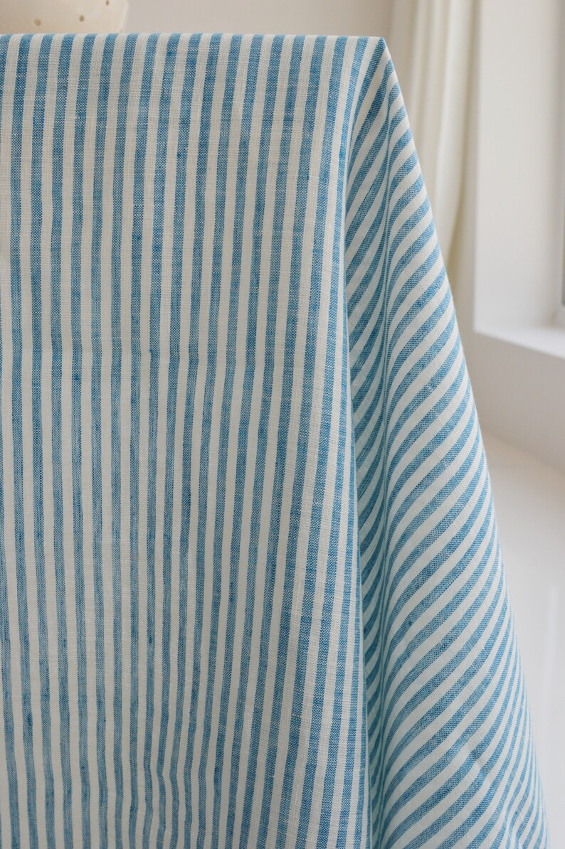 May include: Close-up of a blue and white striped fabric. The fabric is draped, showing vertical stripes of varying widths. The material appears to be linen or a similar natural fiber, with a slightly textured surface. The color palette is a light blue and white.