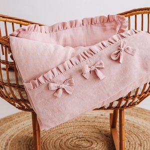 May include: A pink linen baby crib bedding set with ruffled edges and bows. The bedding is in a wicker crib.