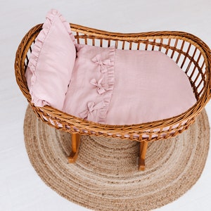 May include: A wicker bassinet with a pink linen bedding set. The bedding includes a pillow and a blanket with ruffled edges and bows.