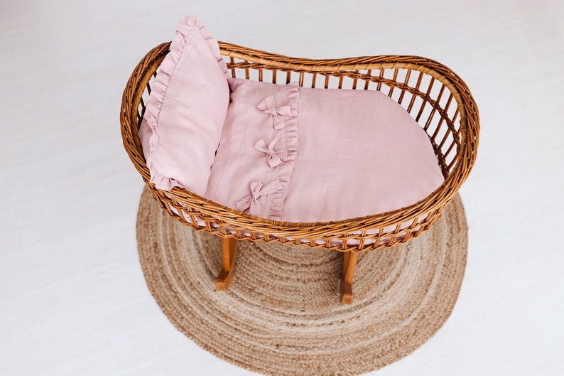 May include: A wicker bassinet with a pink linen bedding set. The bedding includes a pillow and a blanket with ruffled edges and bows.