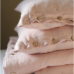 May include: A stack of three pink linen pillowcases with wooden buttons. The pillowcases are folded and arranged on a white surface.