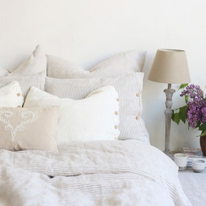 Puede incluir: Una cama blanca con una funda n&oacute;rdica de lino a rayas y varias almohadas. Las almohadas son una mezcla de blanco, beige y gris. Una l&aacute;mpara con una pantalla beige se encuentra en una mesita de noche junto a la cama.