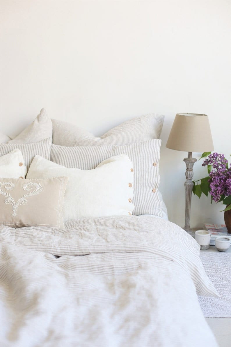 Puede incluir: Una cama blanca con una funda n&oacute;rdica de lino a rayas y varias almohadas. Las almohadas son una mezcla de blanco, beige y gris. Una l&aacute;mpara con una pantalla beige se encuentra en una mesita de noche junto a la cama.