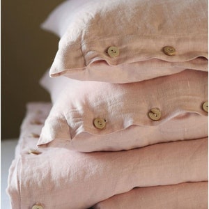 Puede incluir: Una pila de tres fundas de almohada de lino rosa con botones de madera. Las fundas de almohada est&aacute;n dobladas y dispuestas sobre una superficie blanca.