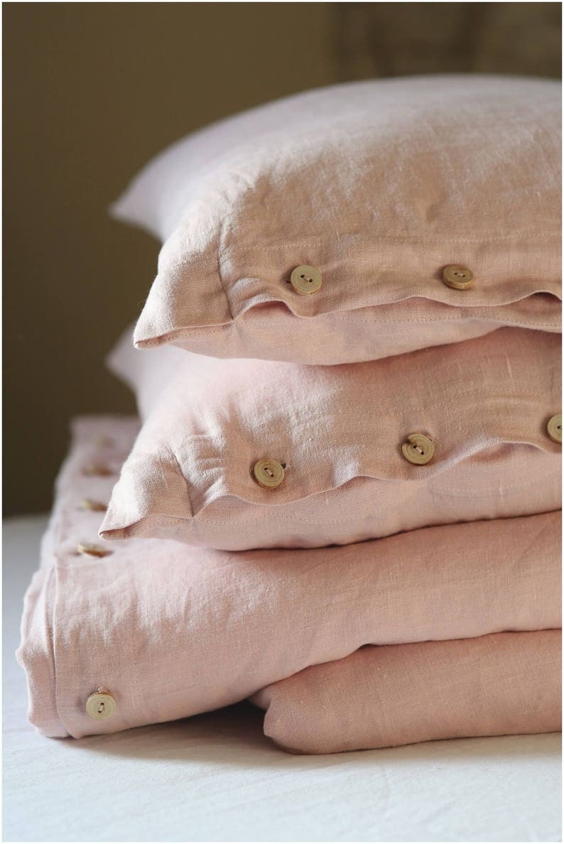 Puede incluir: Una pila de tres fundas de almohada de lino rosa con botones de madera. Las fundas de almohada est&aacute;n dobladas y dispuestas sobre una superficie blanca.