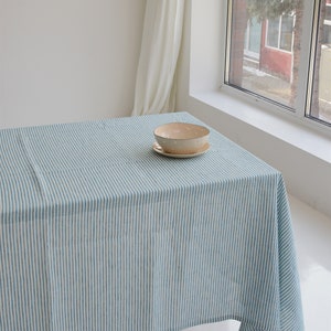 May include: A blue and white striped tablecloth covers a table, with a beige bowl and saucer set on top. The tablecloth has vertical stripes. The scene is lit by natural light from a nearby window.