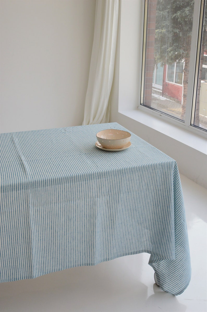 May include: A blue and white striped tablecloth covers a table, with a beige bowl and saucer set on top. The tablecloth has vertical stripes. The scene is lit by natural light from a nearby window.