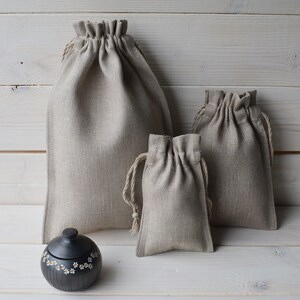 May include: Three beige linen drawstring bags of different sizes on a white wood background. The largest bag is in the back, the medium bag is in the middle, and the smallest bag is in the front. A small black ceramic jar with white daisy designs is in the front of the image.