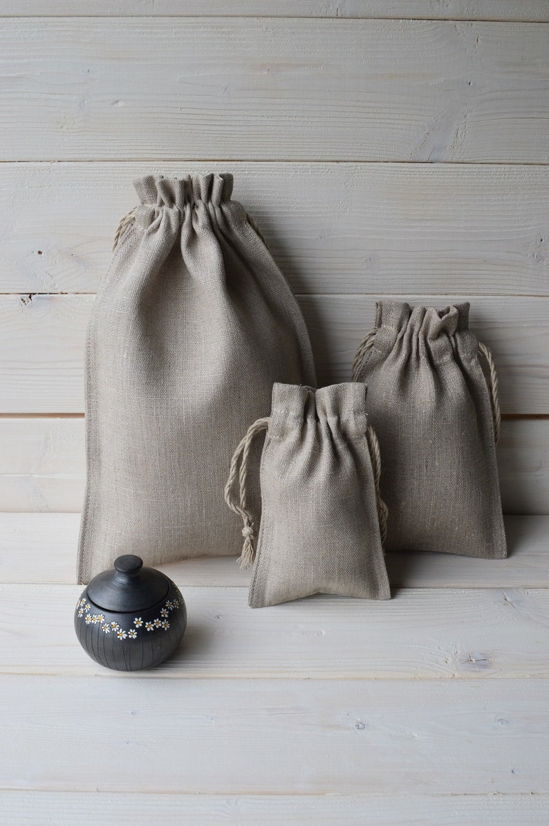 May include: Three beige linen drawstring bags of different sizes on a white wood background. The largest bag is in the back, the medium bag is in the middle, and the smallest bag is in the front. A small black ceramic jar with white daisy designs is in the front of the image.