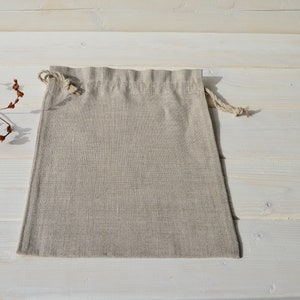 May include: A natural linen drawstring bag with a light beige color. The bag is empty and lying flat on a white wooden surface.