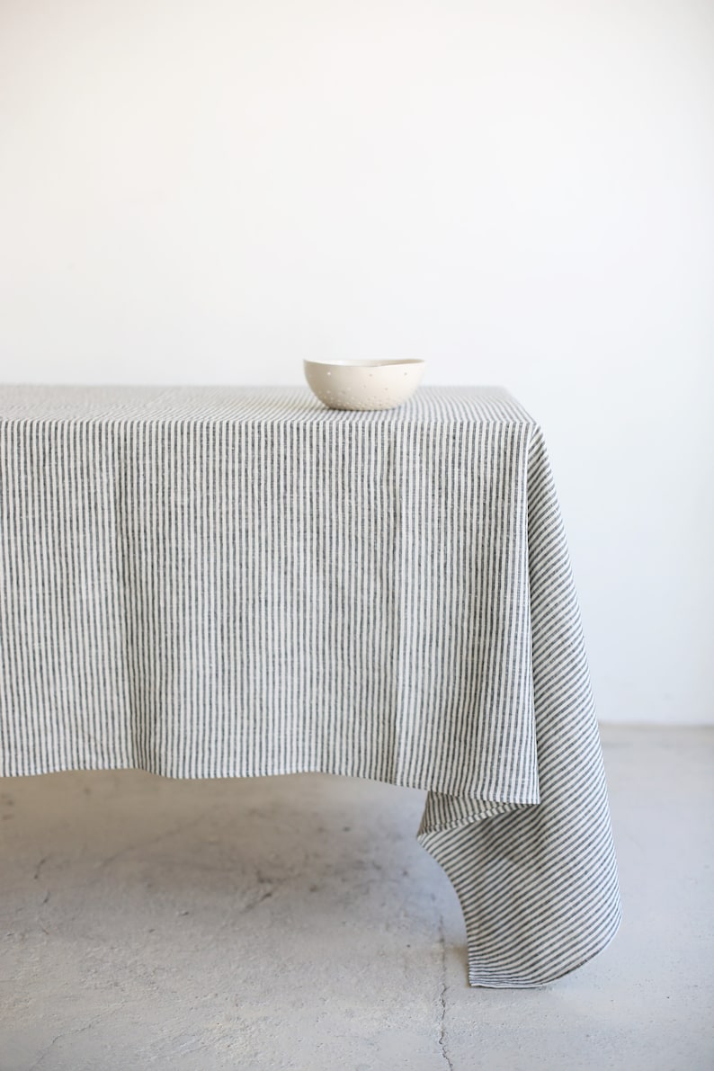 May include: A striped tablecloth in a neutral color palette, featuring vertical lines of dark gray and white. A small, speckled ceramic bowl sits on the table. The tablecloth drapes over the table's edge.