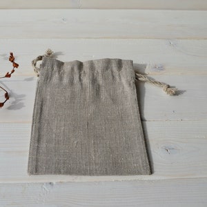 May include: A small, light brown linen drawstring bag with a natural rope drawstring. The bag is lying flat on a white wooden surface.