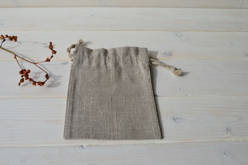 May include: A small, light brown linen drawstring bag with a natural rope drawstring. The bag is lying flat on a white wooden surface.
