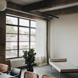 May include: A modern loft interior with a large window, a white wall, a white daybed, a potted plant, and a dining table with chairs.
