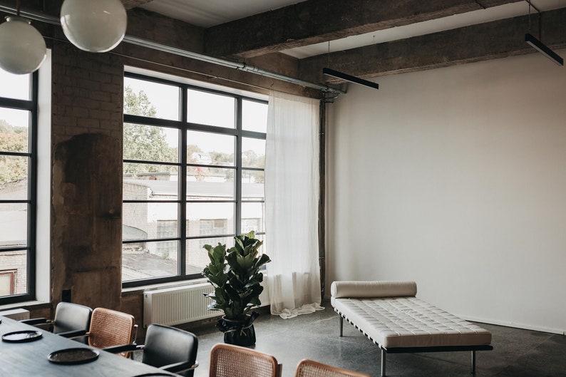 May include: A modern loft interior with a large window, a white wall, a white daybed, a potted plant, and a dining table with chairs.