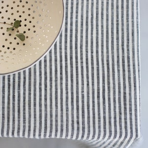 May include: A cream-colored colander with small holes and a sprig of greenery sits on a striped tablecloth. The tablecloth features vertical stripes in shades of gray and white, creating a simple, yet elegant aesthetic.