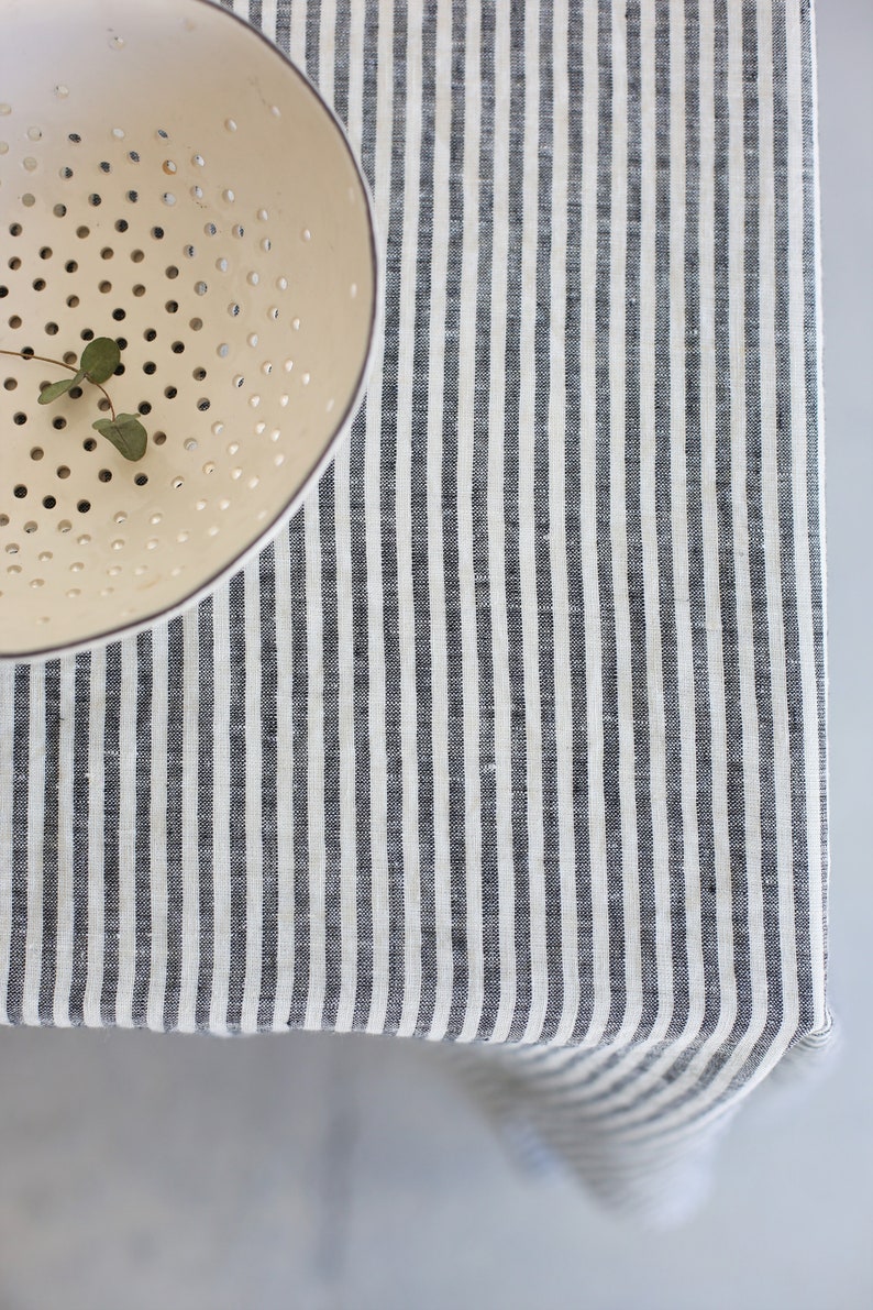 May include: A cream-colored colander with small holes and a sprig of greenery sits on a striped tablecloth. The tablecloth features vertical stripes in shades of gray and white, creating a simple, yet elegant aesthetic.