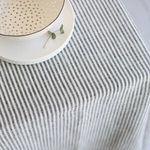 May include: A cream-colored bowl with small holes sits atop a white plate, placed on a striped tablecloth. The tablecloth features vertical stripes in gray and white, creating a simple, neutral aesthetic. A small sprig of greenery rests on the plate.