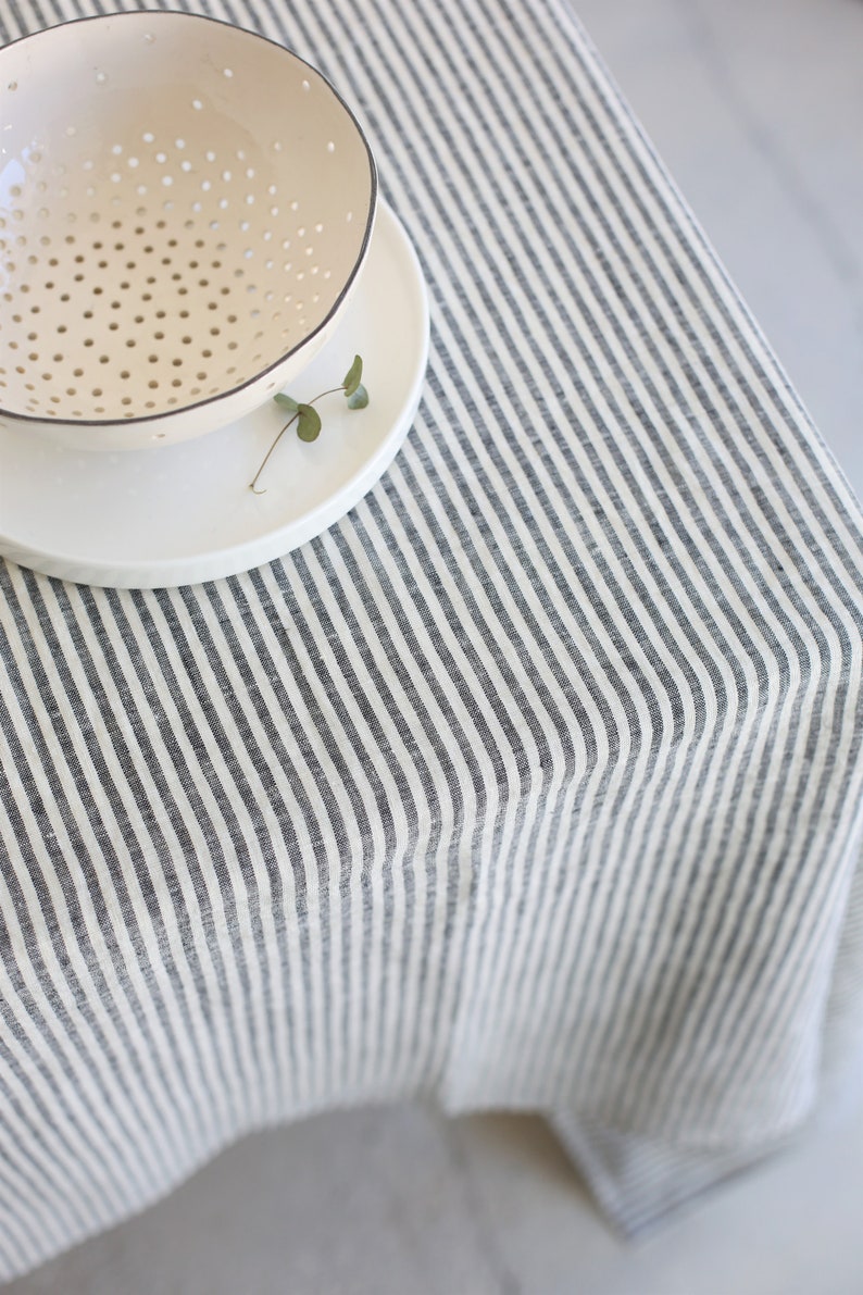 May include: A cream-colored bowl with small holes sits atop a white plate, placed on a striped tablecloth. The tablecloth features vertical stripes in gray and white, creating a simple, neutral aesthetic. A small sprig of greenery rests on the plate.