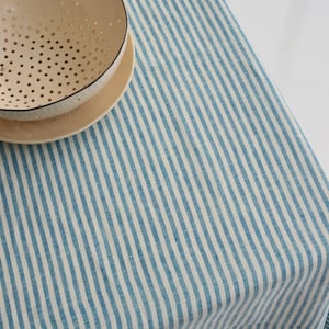 May include: A blue and white striped tablecloth with a beige bowl and plate. The tablecloth has vertical stripes and is made of a linen-like material. The bowl has a perforated design and is sitting on a matching plate.