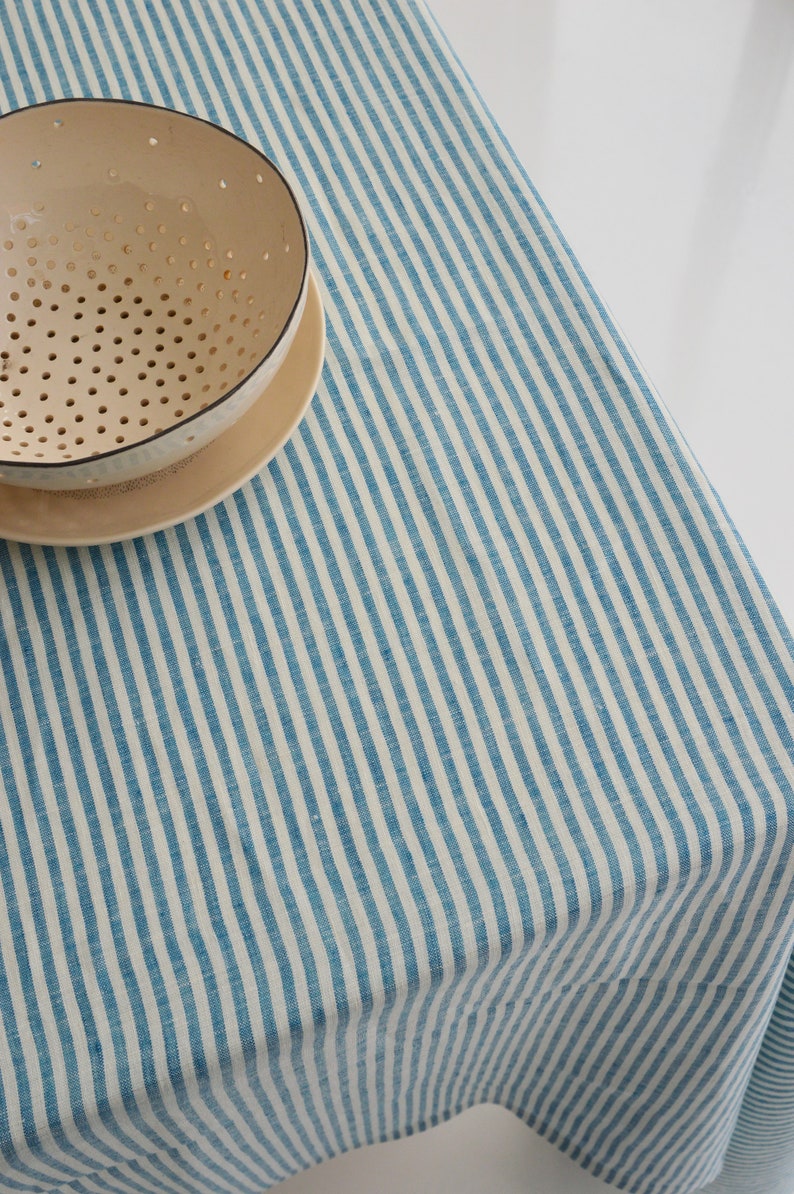 May include: A blue and white striped tablecloth with a beige bowl and plate. The tablecloth has vertical stripes and is made of a linen-like material. The bowl has a perforated design and is sitting on a matching plate.
