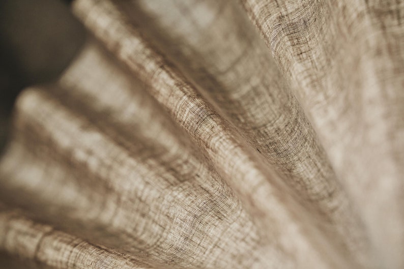 May include: Close-up of a beige fabric with a textured woven pattern. The fabric is folded and draped, creating soft folds and curves.