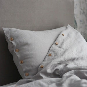 Puede incluir: Una funda de almohada de lino gris con botones de madera. La funda de almohada est&aacute; doblada y los botones son visibles.