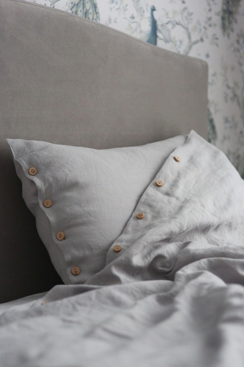 Puede incluir: Una funda de almohada de lino gris con botones de madera. La funda de almohada est&aacute; doblada y los botones son visibles.
