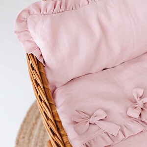 May include: A pink linen baby crib bedding set with a ruffled edge and bows. The bedding is in a wicker basket.