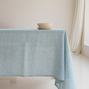 May include: A blue and white striped tablecloth covers a table, with a ceramic bowl and plate set on top. The bowl is beige with small, evenly spaced dots. The tablecloth's pattern is vertical stripes.