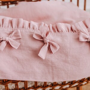 May include: A pink linen baby crib bedding set with a ruffled edge and three bows.