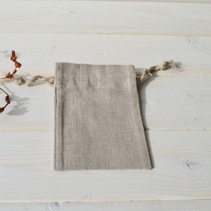 May include: A small, natural linen drawstring bag with a light beige color. The bag is lying flat on a white wooden surface.