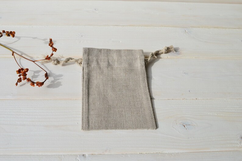May include: A small, natural linen drawstring bag with a light beige color. The bag is lying flat on a white wooden surface.