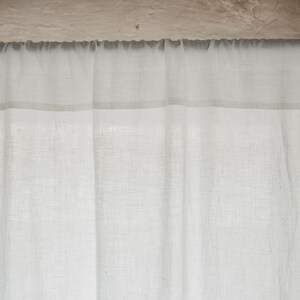 May include: A white sheer curtain with a thin white trim hangs from a metal rod. The curtain is made of a lightweight fabric with a subtle texture.