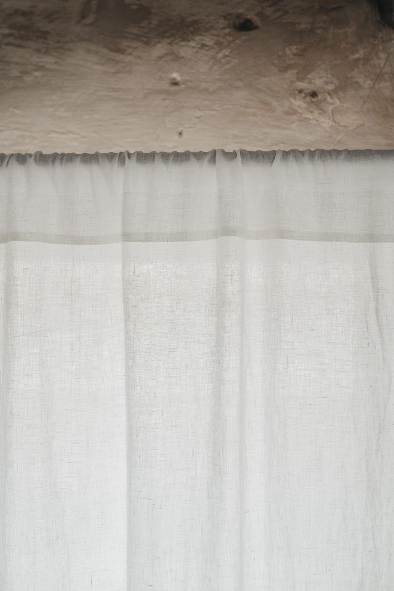 May include: A white sheer curtain with a thin white trim hangs from a metal rod. The curtain is made of a lightweight fabric with a subtle texture.