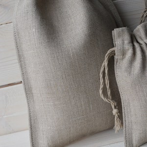 May include: Two natural linen drawstring bags with a light brown color. The bags are made of a woven fabric and have a simple design.