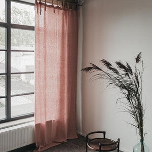 May include: A pink linen curtain hangs in a window with a white radiator below. A brown wicker side table with two shelves sits on a gray tiled floor. A vase with dried grasses sits on the floor next to the table.