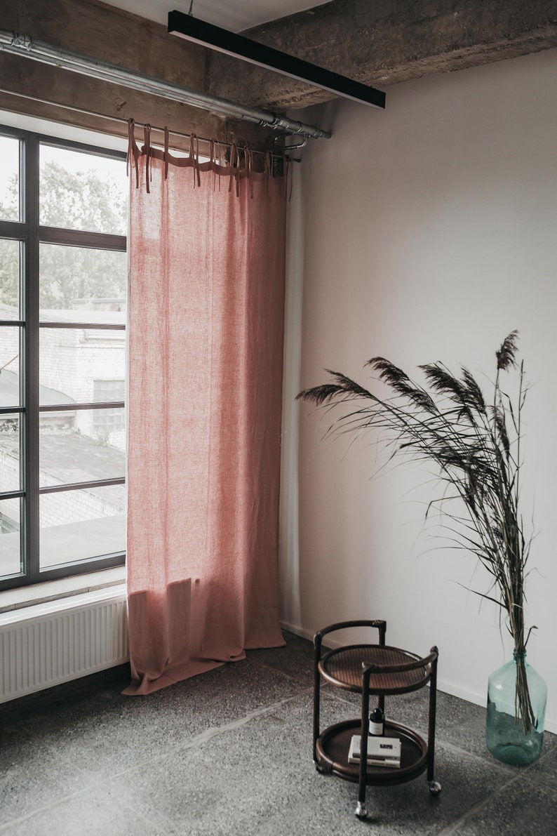 May include: A pink linen curtain hangs in a window with a white radiator below. A brown wicker side table with two shelves sits on a gray tiled floor. A vase with dried grasses sits on the floor next to the table.