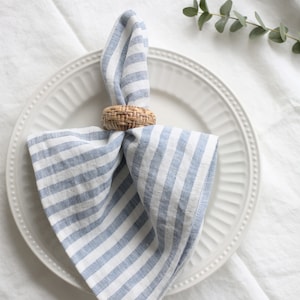 May include: A white plate with a blue and white striped napkin held by a woven napkin ring. The napkin is folded and placed on the plate. The image is a close-up shot of a table setting.
