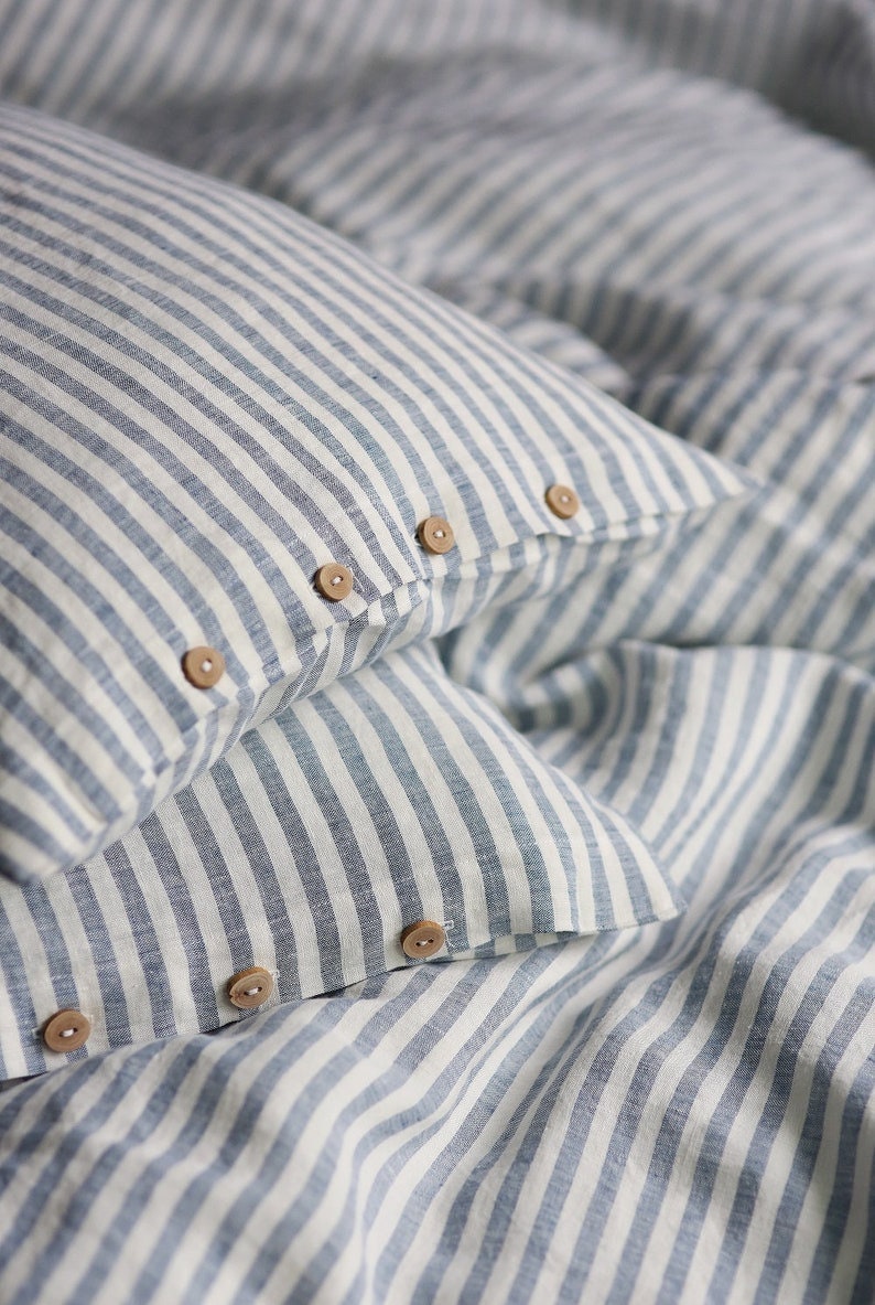 Puede incluir: Dos fundas de almohada de lino a rayas azules y blancas con botones de madera. Las fundas de almohada est&aacute;n dobladas y superpuestas.