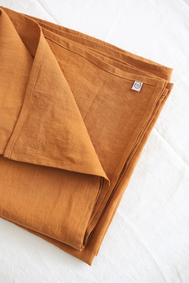 K&ouml;nnte beinhalten: Eine gefaltete, senfgelbe Tischdecke aus Leinen mit einem kleinen wei&szlig;en Etikett, auf dem "Linen & Love" in einem Kreis steht.