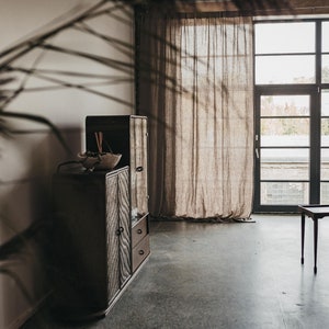 May include: A wooden cabinet with glass doors and drawers, a small table, and a sheer curtain in front of a large window with black metal framing.