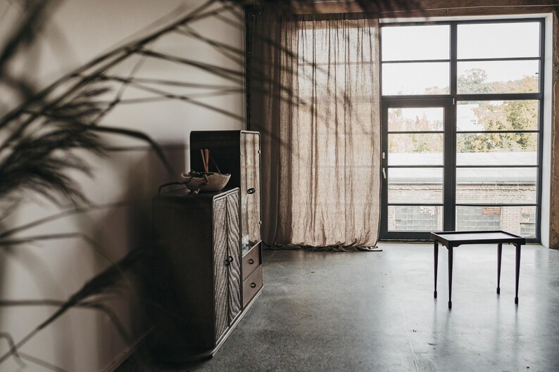 May include: A wooden cabinet with glass doors and drawers, a small table, and a sheer curtain in front of a large window with black metal framing.