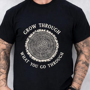 May include: Black t-shirt with a circular tree ring graphic and the text "GROW THROUGH WHAT YOU GO THROUGH" in white. The design features a detailed illustration of tree rings, symbolizing growth and resilience. A casual, everyday wear tee.