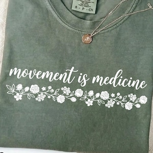 K&ouml;nnte beinhalten: Ein moosgr&uuml;nes T-Shirt mit dem wei&szlig;en Schriftzug "movement is medicine" und einem floralen Design. Eine silberne Halskette mit Anh&auml;nger liegt auf dem T-Shirt, das auf einer neutralen Oberfl&auml;che gefaltet ist.